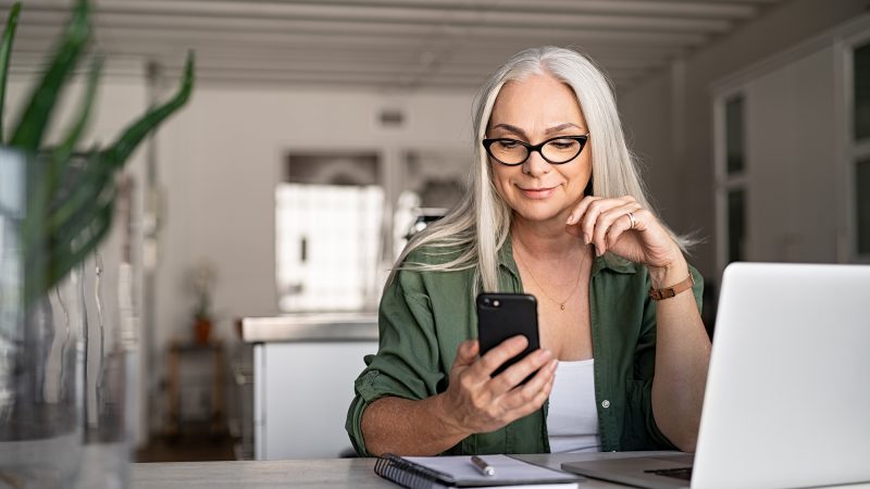 Happy senior woman using mobile phone while working at home with laptop. Smiling cool old woman wearing eyeglasses messaging with smartphone. Beautiful stylish elderly lady browsing site on cellphone.